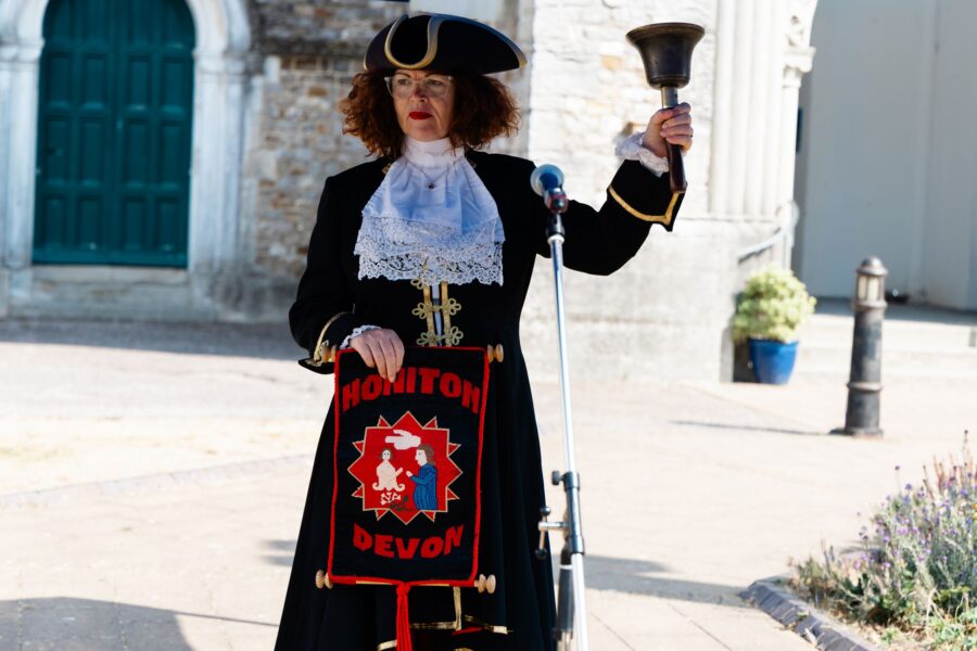 town crier Caroline Kolek ringing a handbell while holding banner which reads "Honiton Devon"