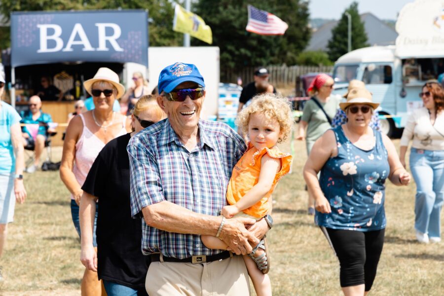 man in baseball cap and sunglasses holding child in arms during line dance at Charter Day 2026