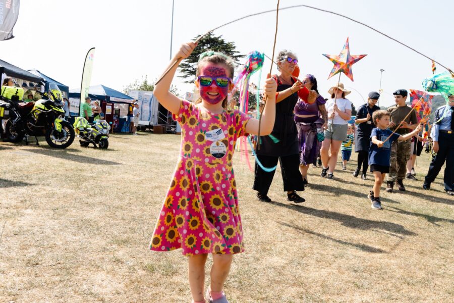 a young girl wearing a pink dress with a sunflower pattern carrying two willow sticks in the children's parade